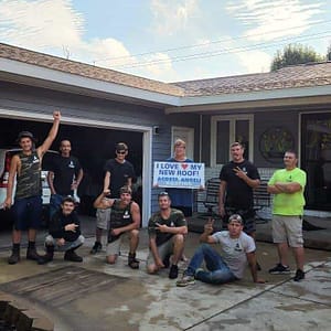 Group of storm damage roofing workers posing proudly in front of a residential garage, with one holding a sign that reads "I love my new roof! - Angela.
